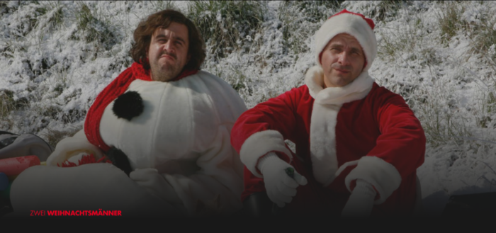 Ausschnitt aus dem Film Zwei Weihnachtsmänner: Zwei Männer - als Weihnachtsmänner verkleidet - sitzen mit ernsten Minen im Schnee. Der Film gehört zu unseren Empfehlungen für Weihnachtsfilme 2018. Foto: Screenshot