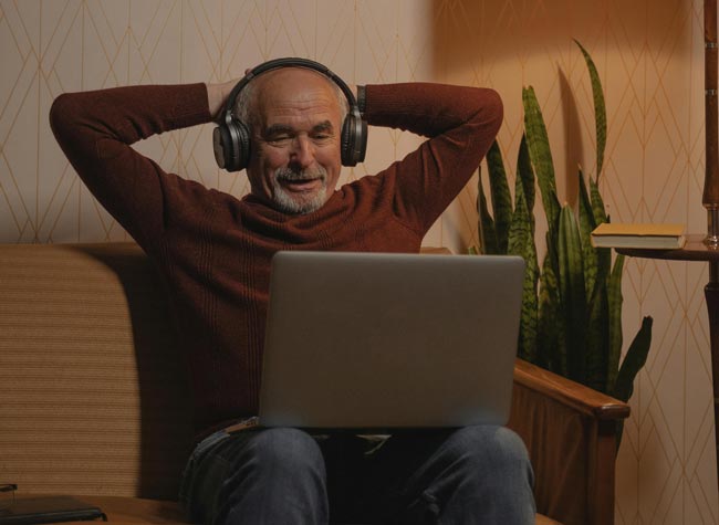 Älterer Mann mit Laptop auf dem Schoß und Kopfhörern, Arme hinterm Kopf verschränkt. Bild: Pexels/Tima Miroshnichenko (https://www.pexels.com/de-de/foto/mann-person-laptop-sitzung-5708715/)