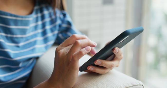 Nahaufnahme einer Frau, die mit den Fingern der rechten Hand auf dem Handy herumtippt, das sie in der linken Hand festhält. Bild: stock.adobe.com/leungchopan (https://stock.adobe.com/de/images/close-up-of-woman-use-of-cellphone-at-home/291863063?asset_id=291863063)