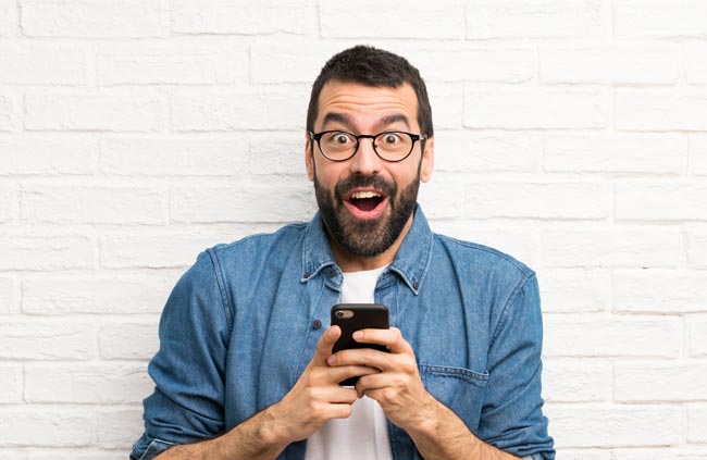Drittanbieter: Freudig blickender Mann mit Brille und Bart, Handy in der Hand. Bild: stock.adobe.com/ luismolinero (https://stock.adobe.com/de/images/handsome-man-with-beard-over-white-brick-wall-surprised-and-sending-a-message/325766606?asset_id=325766606)