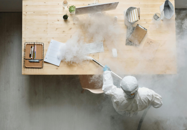Laptop-Tastatur reinigen I Eine Person in Schutzanzug reinigt die Tastatur mit einem Rohr, woraus Nebel kommt. Bild: Pexels/ Michelangelo Buonarroti https://www.pexels.com/de-de/foto/person-tisch-computer-rauch-4176417/