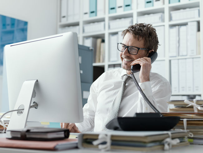 Business-Telefonie | Mann im Büro am Computer mit Telefon am Ohr. Bild: stock.adobe.com/StockPhotoPro 