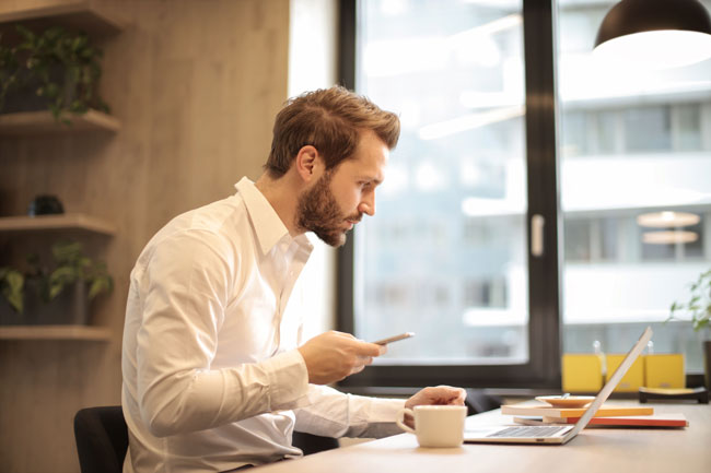 Mann am PC mit Handy checkt E-Mails | Angst vor E-Mails am Arbeitsplatz. Bild: Pexels/Andrea Piacquadio https://www.pexels.com/de-de/foto/mann-im-weissen-hemd-926390/ 
